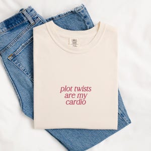 Pode incluir: Camiseta creme com o texto "plot twists are my cardio" em bordô. A camiseta está dobrada e colocada em cima de um par de jeans azuis. A camiseta tem uma etiqueta Comfort Colors.