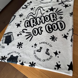 May include: White fabric with black text and illustrations depicting the armor of God. The text includes "Armor of God", "The Gospel of Peace", "Breastplate of", "Belt of Truth", "Sword of the Spirit", "Helmet of Salvation", "Sandals of the Gospel of Peace".