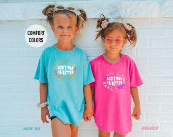 May include: Two young girls wearing matching t-shirts with the text "God's Way Is Better". One shirt is lagoon blue, the other is crunchberry. The shirts have a round neck and short sleeves. The girls are standing in front of a white brick wall.