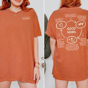 May include: A rust-colored t-shirt with a white graphic design on the back. The graphic features a circle with the words "Good News" inside. The circle is surrounded by arrows and other symbols, and includes text about God's love and sacrifice.