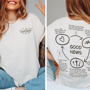 May include: White t-shirt with a graphic design of a heart, cross, and the words "Good News". The back of the shirt has a diagram with the words "Good News" and text about God's love, Jesus' sacrifice, and the free gift of eternal life.