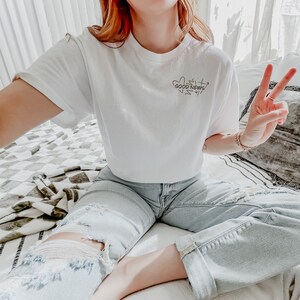 May include: A white cropped t-shirt with the text "Good News" in a handwritten style. The shirt is worn with light blue distressed jeans.
