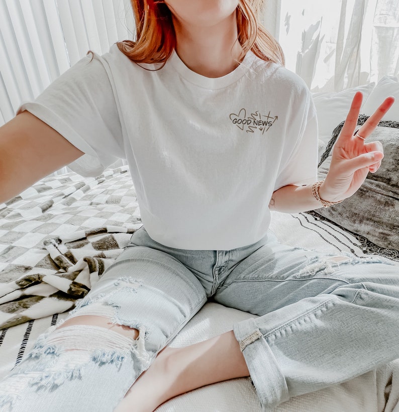 May include: A white cropped t-shirt with the text "Good News" in a handwritten style. The shirt is worn with light blue distressed jeans.
