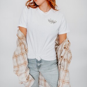 May include: A white t-shirt with a black graphic that says "Good News" with a heart and arrows around the text. The model is wearing the shirt with a plaid shirt over it and blue jeans.