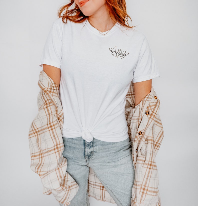 May include: A white t-shirt with a black graphic that says "Good News" with a heart and arrows around the text. The model is wearing the shirt with a plaid shirt over it and blue jeans.