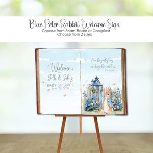 May include: A Peter Rabbit-themed welcome sign for a baby shower. The sign features a blue and white design with the text "Welcome to Beth & Jake's BABY SHOWER June 29, 2024." The sign is displayed on a wooden easel.