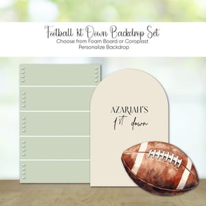 May include: A beige backdrop with an arch shape and the text "AZARIAH'S 1st down" in black. A brown and white football is in the bottom right corner of the backdrop.