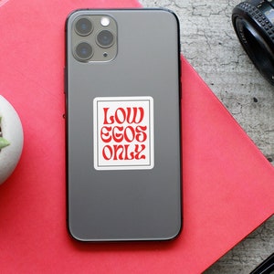 May include: A grey iPhone with a white sticker that says "LOW EGOS ONLY" in red letters.
