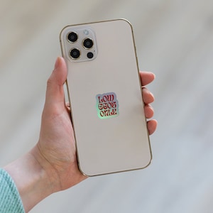 May include: A white iPhone with a gold trim and a camera bump. The phone has a sticker on the back that says "LOW  EGOS ONLY" in red letters.