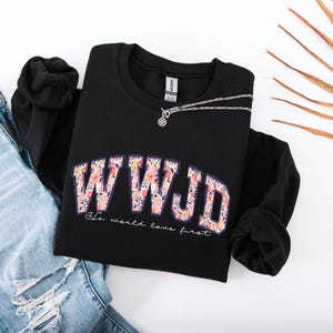 May include: Black sweatshirt with the text "WWJD" in a floral design. The phrase "He would love first" is printed below the main text. A silver necklace is draped across the sweatshirt, which is laid on top of a pair of blue jeans.