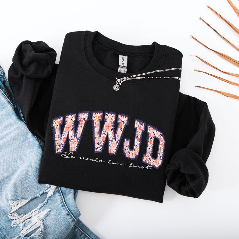May include: Black sweatshirt with the text "WWJD" in a floral design. The phrase "He would love first" is printed below the main text. A silver necklace is draped across the sweatshirt, which is laid on top of a pair of blue jeans.