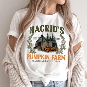 May include: White t-shirt featuring a graphic design with the text "Hagrid's Pumpkin Farm" in orange and green. The design includes an illustration of a cottage, pumpkins, and the text "Magically Grown."