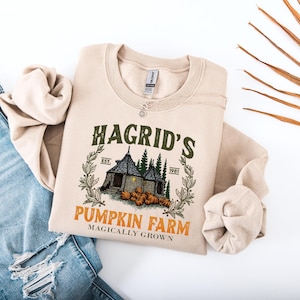 Peut inclure: Sweat-shirt beige avec l'inscription "Hagrid's Pumpkin Farm" et une illustration d'un cottage, de citrouilles et d'arbres. Le texte "Est. 1981" est également inclus. Le sweat-shirt est plié sur une surface blanche avec un jean bleu.