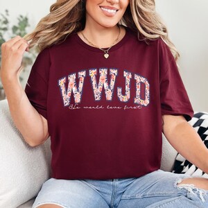 May include: A maroon crew neck t-shirt with the text "WWJD" in a floral design. Below the text is the phrase "He would love first". The shirt is a casual style.