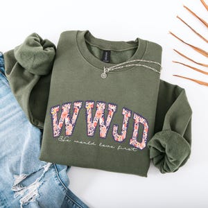 May include: An olive green sweatshirt with the letters "WWJD" in a floral design. Below the letters, the text reads "He would love first." A silver necklace and blue jeans are also in the image.
