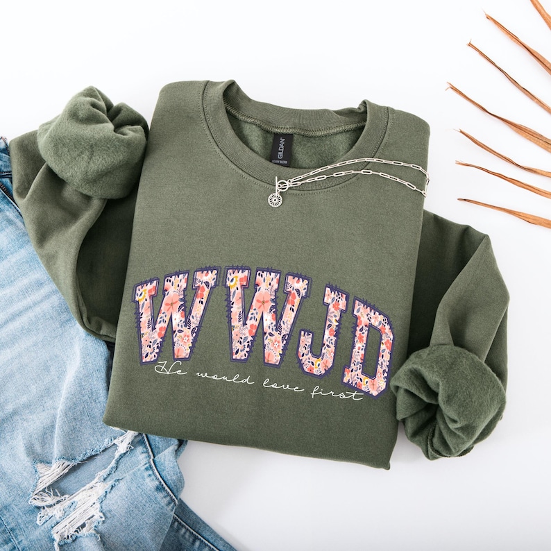 May include: An olive green sweatshirt with the letters "WWJD" in a floral design. Below the letters, the text reads "He would love first." A silver necklace and blue jeans are also in the image.
