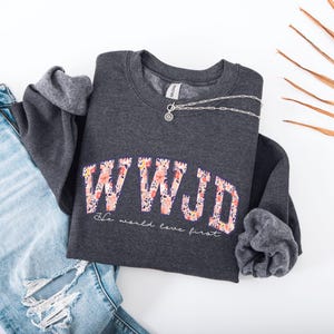 May include: A dark gray sweatshirt with the letters "WWJD" in a floral design. Below the letters, the text reads "He would love first." A silver necklace and blue jeans are also in the image.