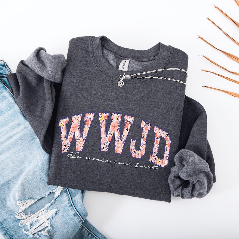 May include: A dark gray sweatshirt with the letters "WWJD" in a floral design. Below the letters, the text reads "He would love first." A silver necklace and blue jeans are also in the image.