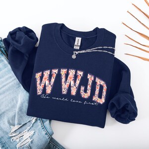 May include: A navy blue sweatshirt with the letters "WWJD" in a floral design. The text "He would love first" is below the letters. A silver necklace is draped across the sweatshirt, which is placed on top of a pair of blue jeans.
