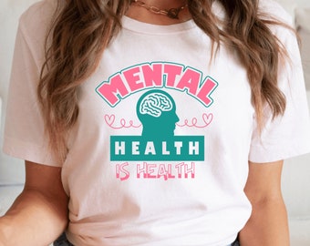 May include: White t-shirt with a pink and teal graphic that says "MENTAL HEALTH IS HEALTH". The graphic includes a teal brain and profile silhouette.
