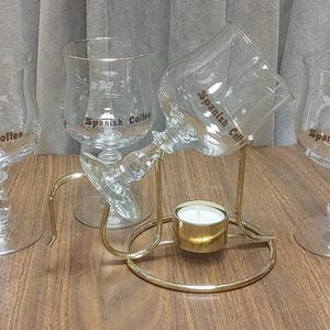 Op de afbeelding: Vier heldere glazen koffiemokken met de tekst "Spanish Coffee" erop gedrukt. Een goudkleurige metalen standaard houdt een van de glazen en een kleine kaars vast. De glazen staan op een houten oppervlak.