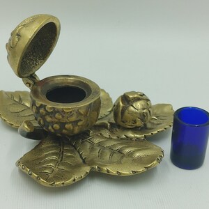 May include: A brass inkwell shaped like an acorn with a lid, sitting on a brass leaf design. A small blue glass inkwell sits to the right.