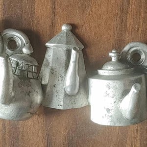 May include: Five silver-coloured teapot ornaments with varying designs, including scalloped edges and geometric shapes. The teapots are arranged on a wooden surface, showcasing their metallic finish and intricate details.