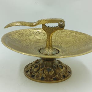 May include: A gold-toned metal nutcracker with a decorative base and a small dish. The nutcracker has a lever and a hinged jaw. The base is ornate and has a floral design.