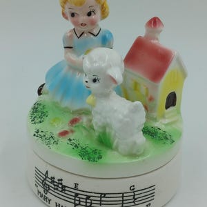 May include: A porcelain musical trinket box featuring a girl in a blue dress holding a white lamb. The box is decorated with a yellow and red house and green grass. The bottom of the box has the words "Mary Had A Little Lamb" and musical notes.