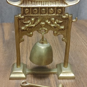 May include: A small, brass-colored bell with a pagoda-style frame. The bell hangs from a decorative archway supported by two pillars. A small mallet rests on the surface. The bell and frame are made of a shiny, metallic material.