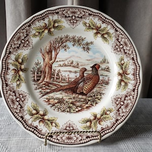 Op de afbeelding: Een decoratieve vintage bord met een fazantenontwerp. Het bord heeft een bruin-witte rand met bloemen en eikels. Het midden toont twee fazanten in een landschap met een boom, hek en heuvels. Het bord staat op een goudkleurige standaard.