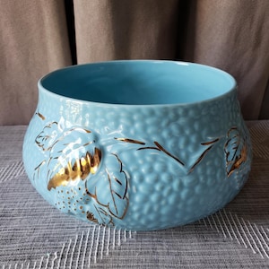 May include: A light blue ceramic bowl with a textured, pebbled surface. The exterior features embossed leaf and vine patterns highlighted with metallic gold paint. The bowl has a wide, open circular top and a rounded, bulbous body shape.