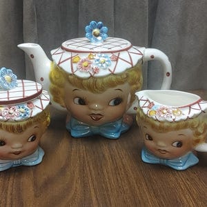 Op de afbeelding: Een vintage keramische theeset met een theepot, suikerpot en melkkan. Elk stuk is ontworpen met een grillig gezicht, blond haar en een blauwe vlinderdas. De theepot heeft een bloemen deksel en de melkkan heeft een handvat.