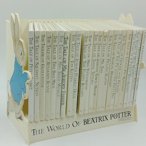 Peut inclure: Une bibliothèque blanche avec une découpe de Lapin de Pâques sur le devant. La bibliothèque est remplie de 24 livres, tous portant le titre "L'Histoire de..." et l'auteur Beatrix Potter. Les livres sont disposés dans l'ordre de gauche à droite, avec les titres visibles sur les dos.