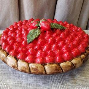 May include: A ceramic strawberry pie plate with a brown crust and a glossy red strawberry topping. The pie is decorated with green leaves and additional strawberries.
