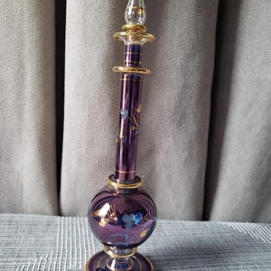 Peut inclure: Un flacon de parfum en verre violet avec un long col fin et une base sphérique. Le flacon présente des bordures dorées à la base, au col et sur le bouchon, ainsi que de délicats motifs floraux peints en doré et bleu clair sur le verre.