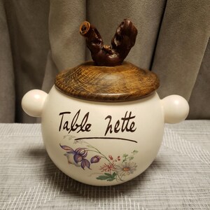 May include: A white ceramic container with a wooden lid and a natural wood handle. The container has two small handles on the sides and features the text "Table nette" with floral illustrations.