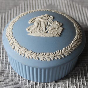 May include: A round, light blue ceramic trinket box with a matte finish. The lid features a white relief of classical figures and a surrounding laurel leaf border. The sides of the box have a vertical ribbed texture, creating a consistent design.