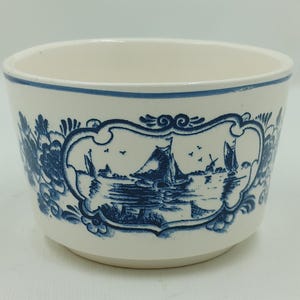 May include: A white ceramic bowl with a blue and white design featuring a Dutch seascape with sailboats and a windmill. The bowl has a scalloped edge and a floral pattern around the rim.