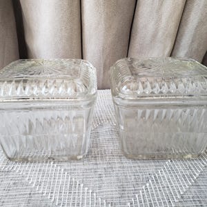 以下が含まれることがあります： 蓋付きの透明なガラス製の正方形の容器が2つ。容器の側面と蓋には、テクスチャ加工された幾何学模様が施されています。蓋の中央には、装飾的なスターバースト模様があります。容器は白とグレーの模様の表面に置かれています。