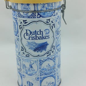 Könnte beinhalten: Eine vintage Blechdose in Blau und Weiß mit einem gelben Deckel. Die Dose hat ein holländisches Thema mit Bildern von Windmühlen, Pferden und anderen holländischen Szenen. Der Text "Dutch Crisbakes" ist auf der Vorderseite der Dose aufgedruckt.