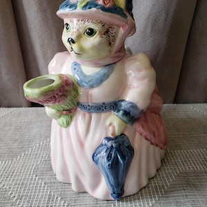 May include: A ceramic bear figurine wearing a pink dress and hat, holding a bouquet and a blue umbrella. The bear has a light brown face and a hat with floral details. This decorative figurine is a charming collectable item.