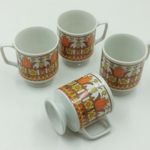 May include: Four white ceramic mugs with a retro floral design featuring orange tulips and yellow flowers. The mugs have a brown fence pattern and a handle. The mugs are stacked on a white surface.