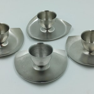 May include: Four silver egg cups with matching saucers. Each cup is made of polished metal and has a rounded shape. The saucers are slightly curved and have a smooth surface.