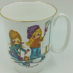 May include: White ceramic mug with a gold rim. The mug features a colourful illustration of two girls painting. The text "Give a helping hand" is printed on the bottom of the mug.