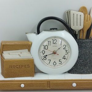 May include: White kitchen clock with a teapot design, featuring a black clock face with black hands and Roman numerals. The clock is mounted on a wooden shelf with a recipe box and a utensil holder with a grey finish.