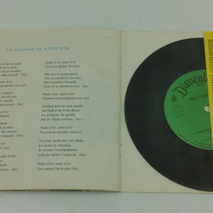 May include: An open book with French lyrics and a black vinyl record. The record has a green label with the Disney logo. A yellow paper with text is on the right side.