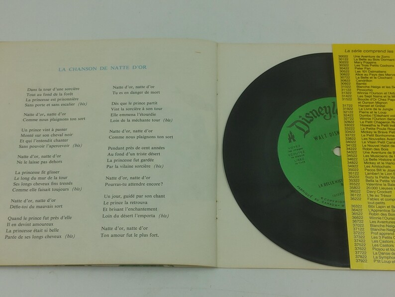 May include: An open book with French lyrics and a black vinyl record. The record has a green label with the Disney logo. A yellow paper with text is on the right side.