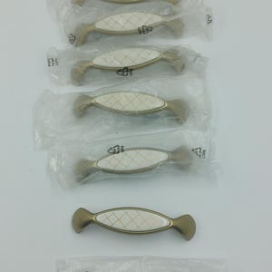 May include: Six gold cabinet knobs with white ceramic inserts and a diamond pattern. The knobs are packaged in individual plastic bags. A seventh knob is shown separately with a bag of screws.
