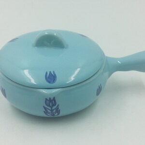 May include: A pale blue ceramic casserole dish with a lid and handle. The dish is decorated with dark blue floral designs. The handle is on the right side. The dish is designed for cooking and serving, with a capacity of approximately 250 ml.